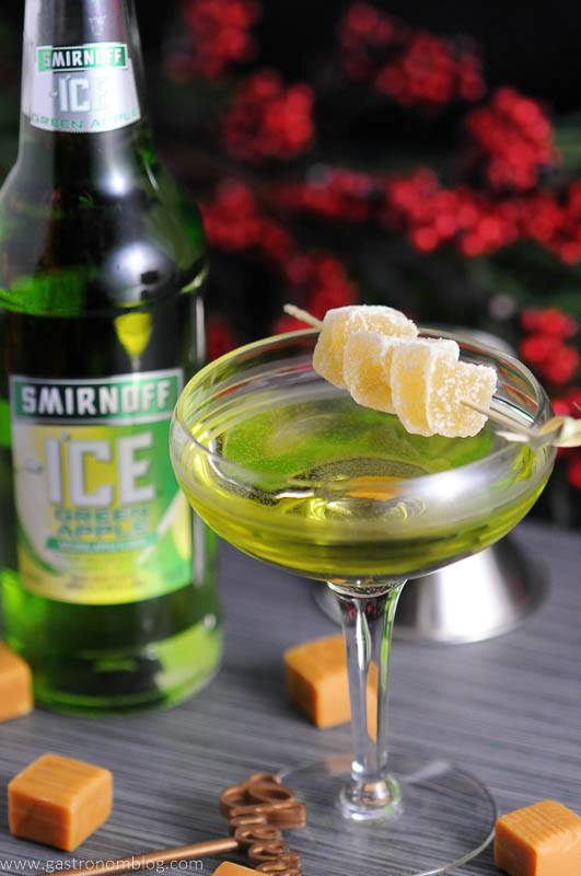A green cocktail in a martini glass, garnished with candied ginger, sits beside a bottle of Smirnoff Ice Green Apple, with caramel candies and a jigger nearby.