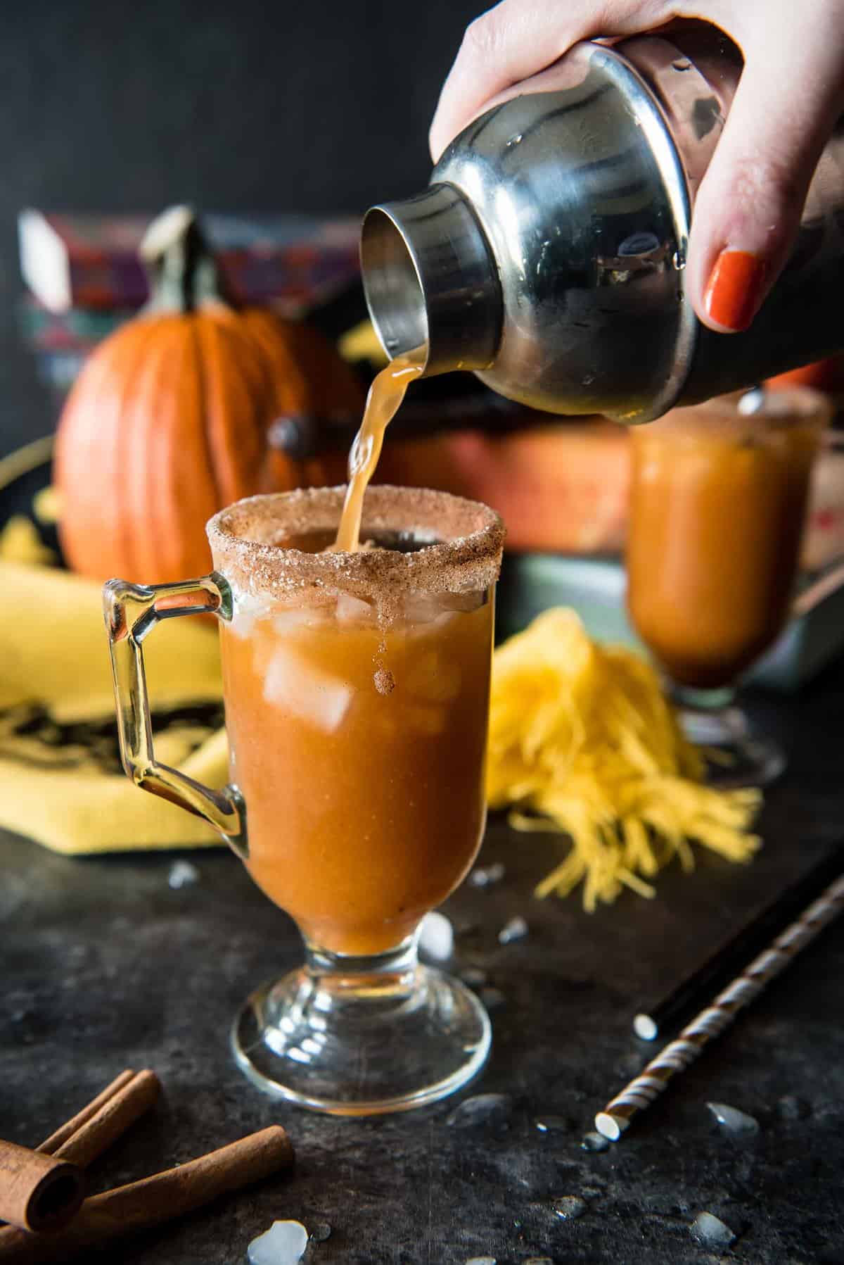 A hand pours an orange-brown drink from a cocktail shaker into a sugar-rimmed glass with ice. A pumpkin and yellow cloth are in the background, with cinnamon sticks nearby.