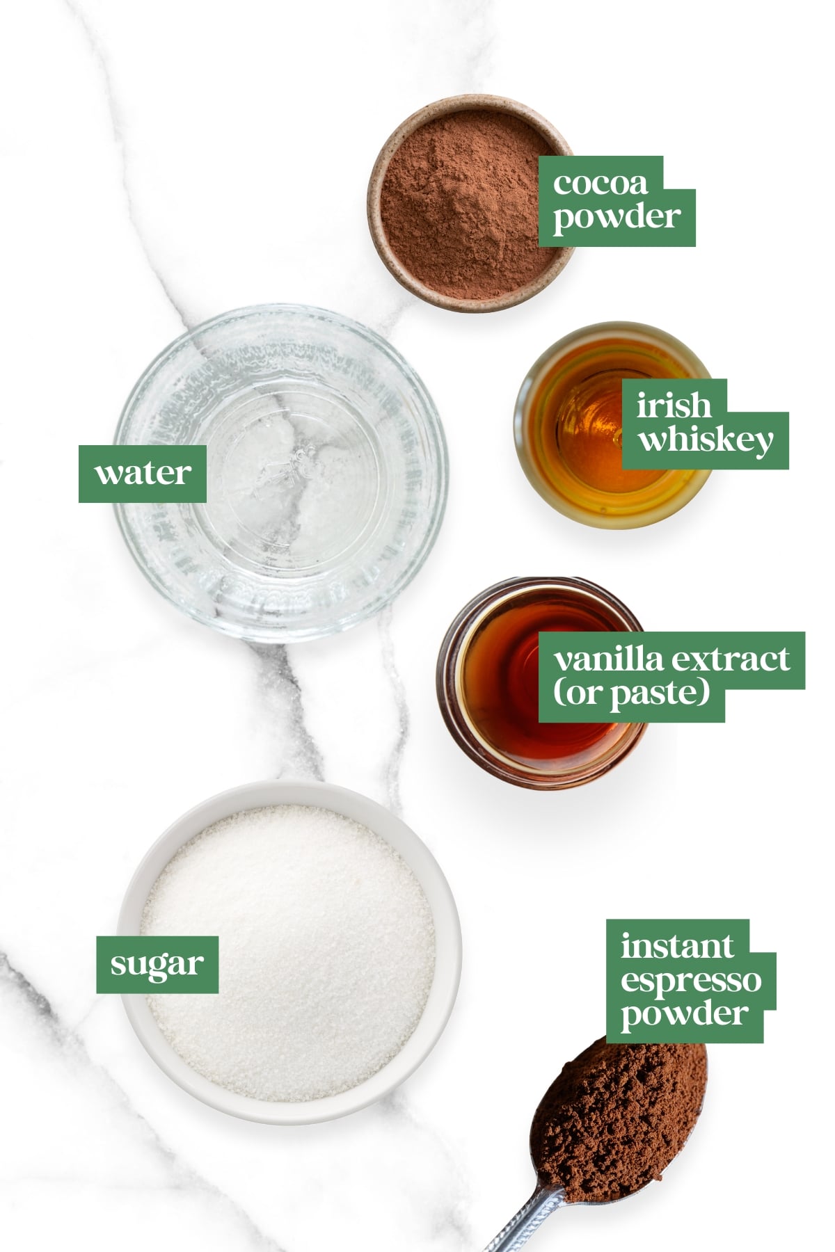 Five ingredients for how to make Irish coffee are labeled on a marble surface: cocoa powder, water, Irish whiskey, vanilla extract (or paste), sugar, and instant espresso powder.