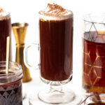A glass mug of dark hot chocolate topped with whipped cream and cocoa powder sits on a white surface, surrounded by a jar, a carafe, and a measuring jigger.
