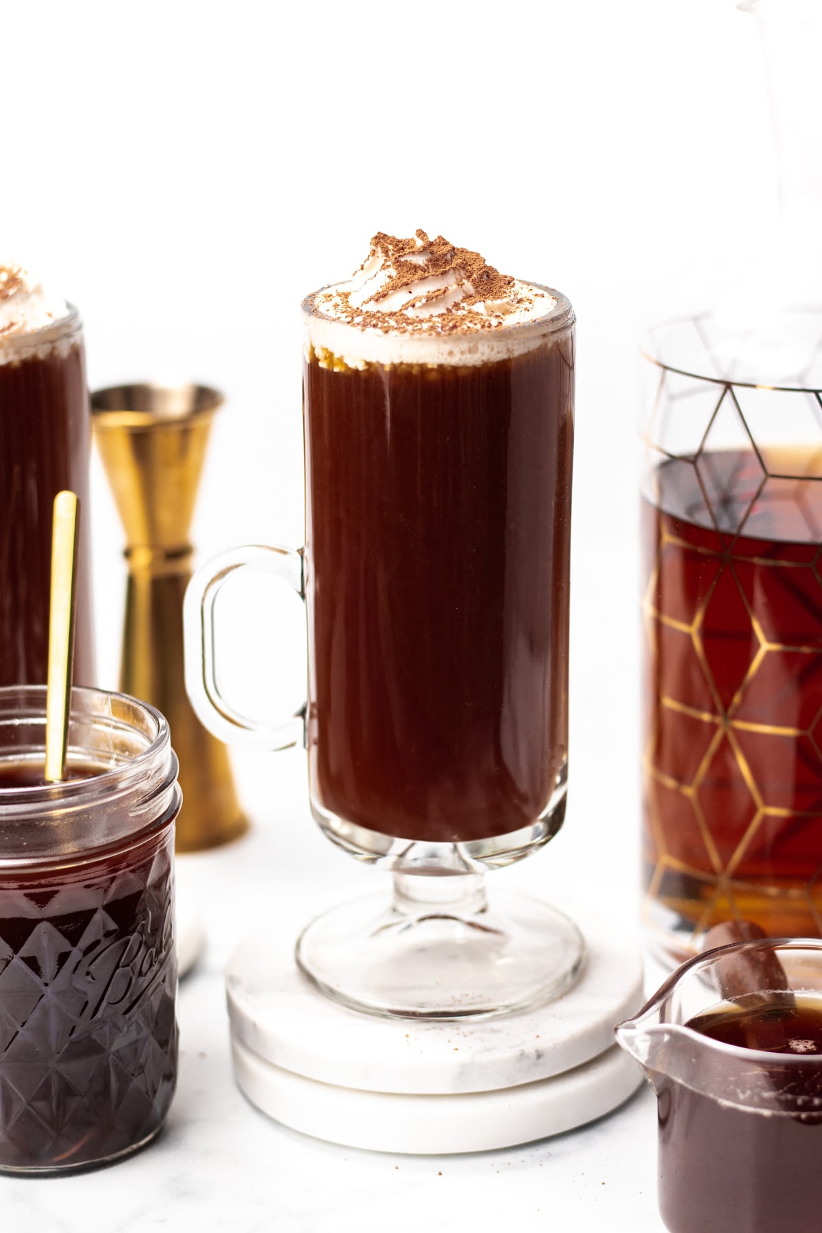 A glass mug of dark hot chocolate topped with whipped cream and cocoa powder sits on a white surface, surrounded by a jar, a carafe, and a measuring jigger.