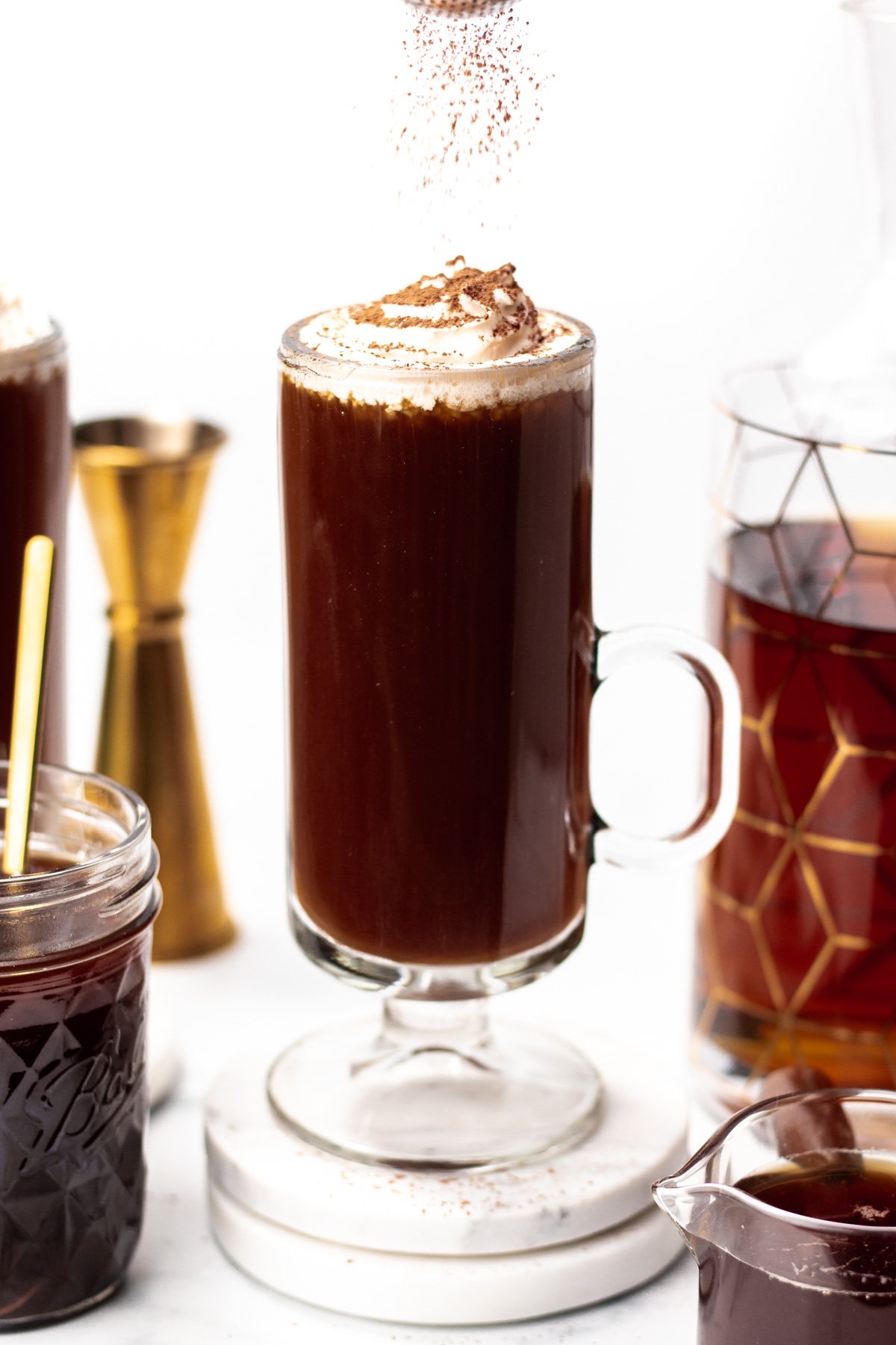A glass mug of dark coffee topped with whipped cream and chocolate shavings, surrounded by a jar, a jigger, and a decanter of dark liquid on a white surface.