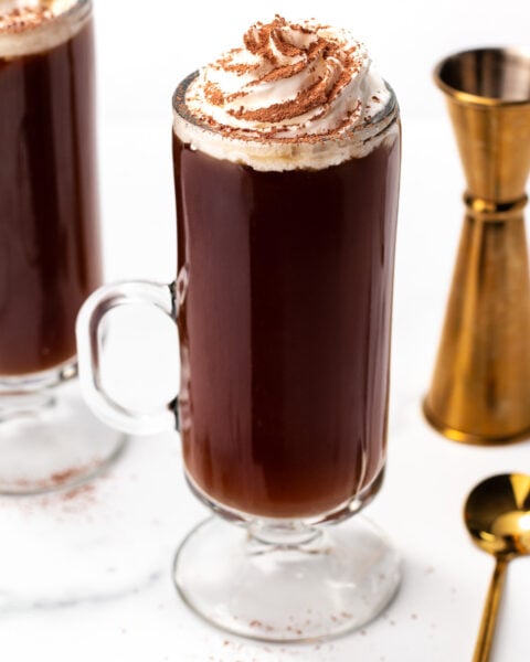 A glass mug of dark coffee topped with whipped cream and chocolate shavings, next to a gold jigger and spoon on a white surface.