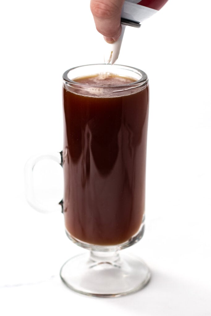 A hand pours creamer from a small container into a glass mug filled with black coffee.