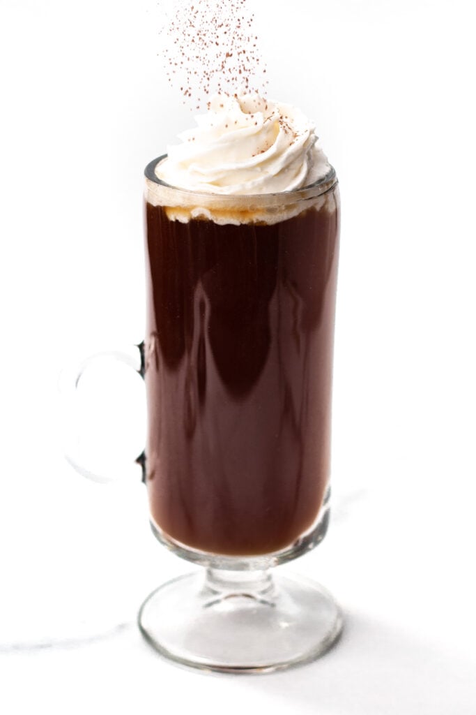 A glass mug of dark coffee topped with whipped cream and sprinkled with cocoa powder, set against a white background.