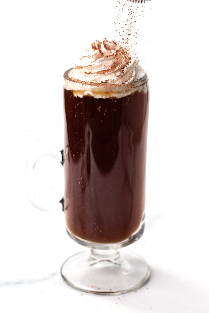 A glass mug filled with dark coffee, topped with a swirl of whipped cream and a dusting of cocoa powder, against a white background.