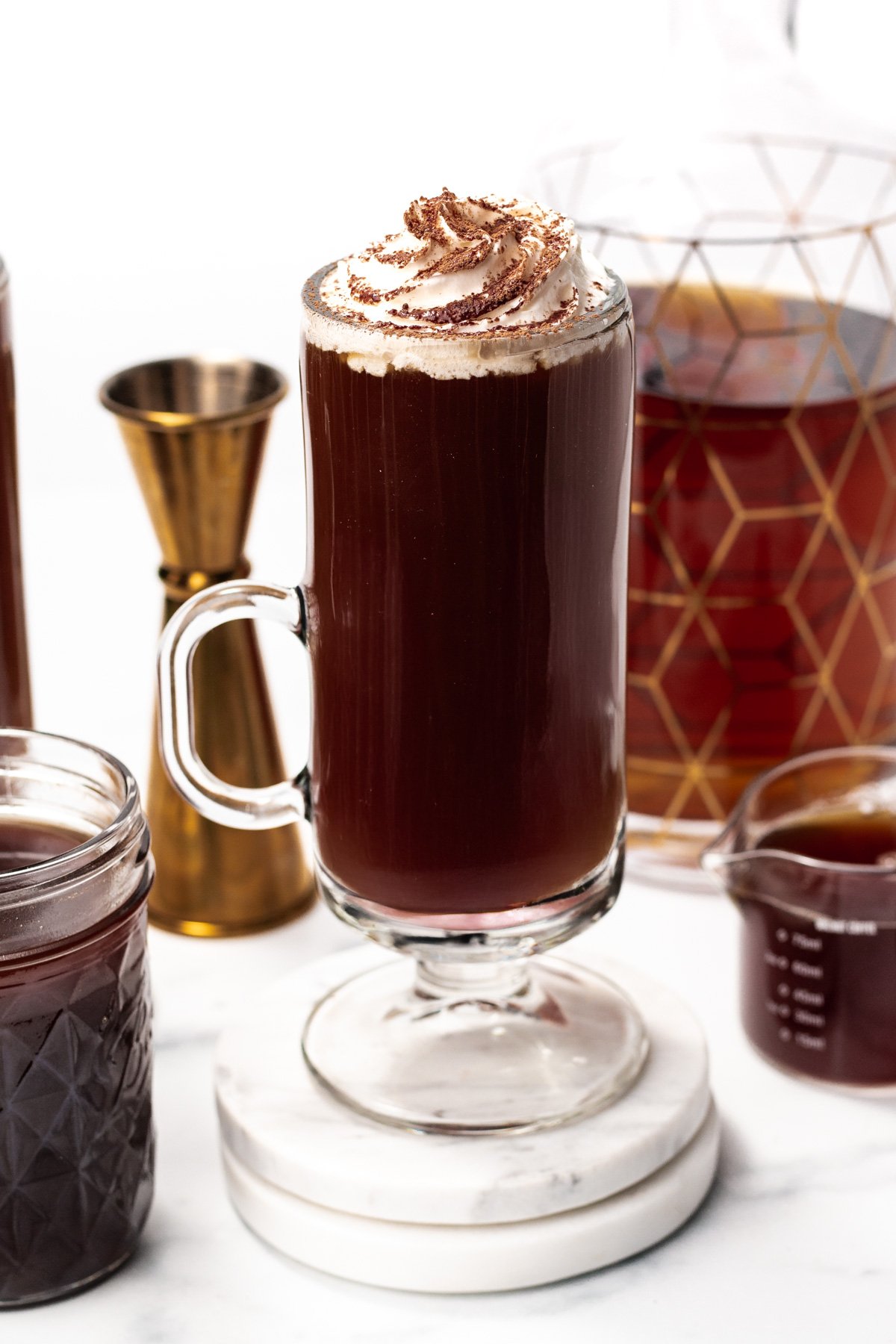 A glass mug of dark coffee topped with whipped cream and chocolate shavings, surrounded by a jar, a jigger, a decanter, and a measuring cup filled with liquid.