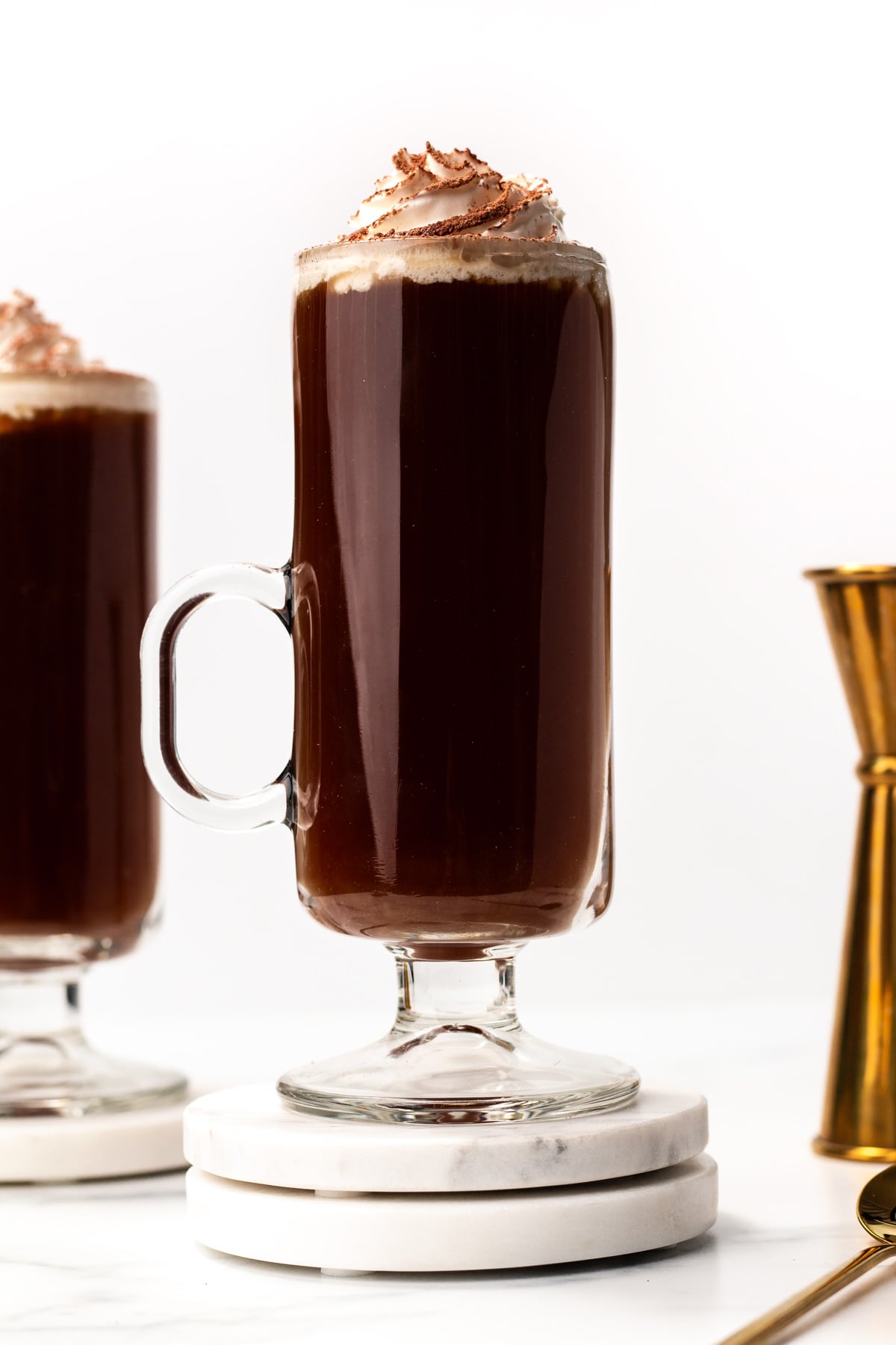A tall glass mug filled with dark coffee topped with whipped cream and chocolate shavings, placed on a white coaster. Another mug and a gold jigger are visible in the background.