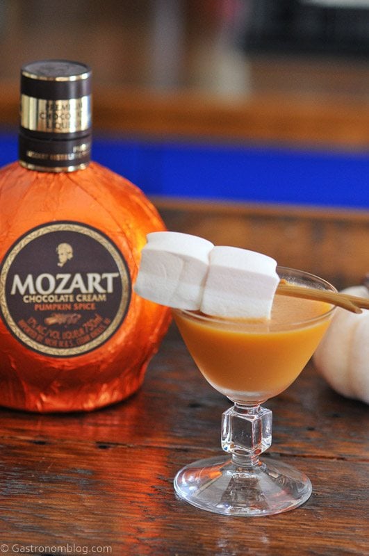 A glass of brown cocktail garnished with three marshmallows on a skewer, next to a bottle of Mozart Chocolate Cream Pumpkin Spice liqueur.