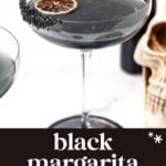 A black margarita cocktail in a coupe glass, garnished with black salt and a dried citrus slice, with a skull decoration in the background.