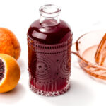 A glass bottle filled with dark red juice is placed between a halved blood orange, a whole blood orange, and a pink glass citrus juicer on a white surface.