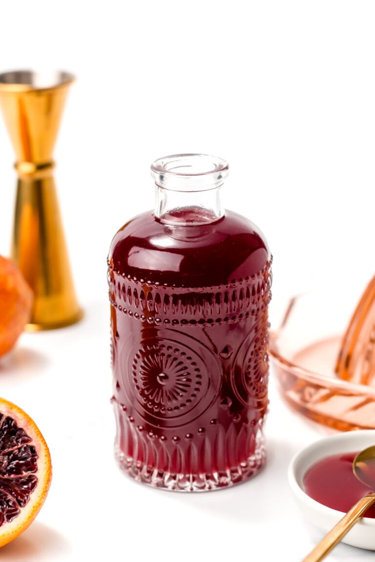 A decorative glass bottle filled with dark red liquid sits on a white surface, surrounded by a halved blood orange, a brass jigger, a citrus juicer, and a bowl of juice.