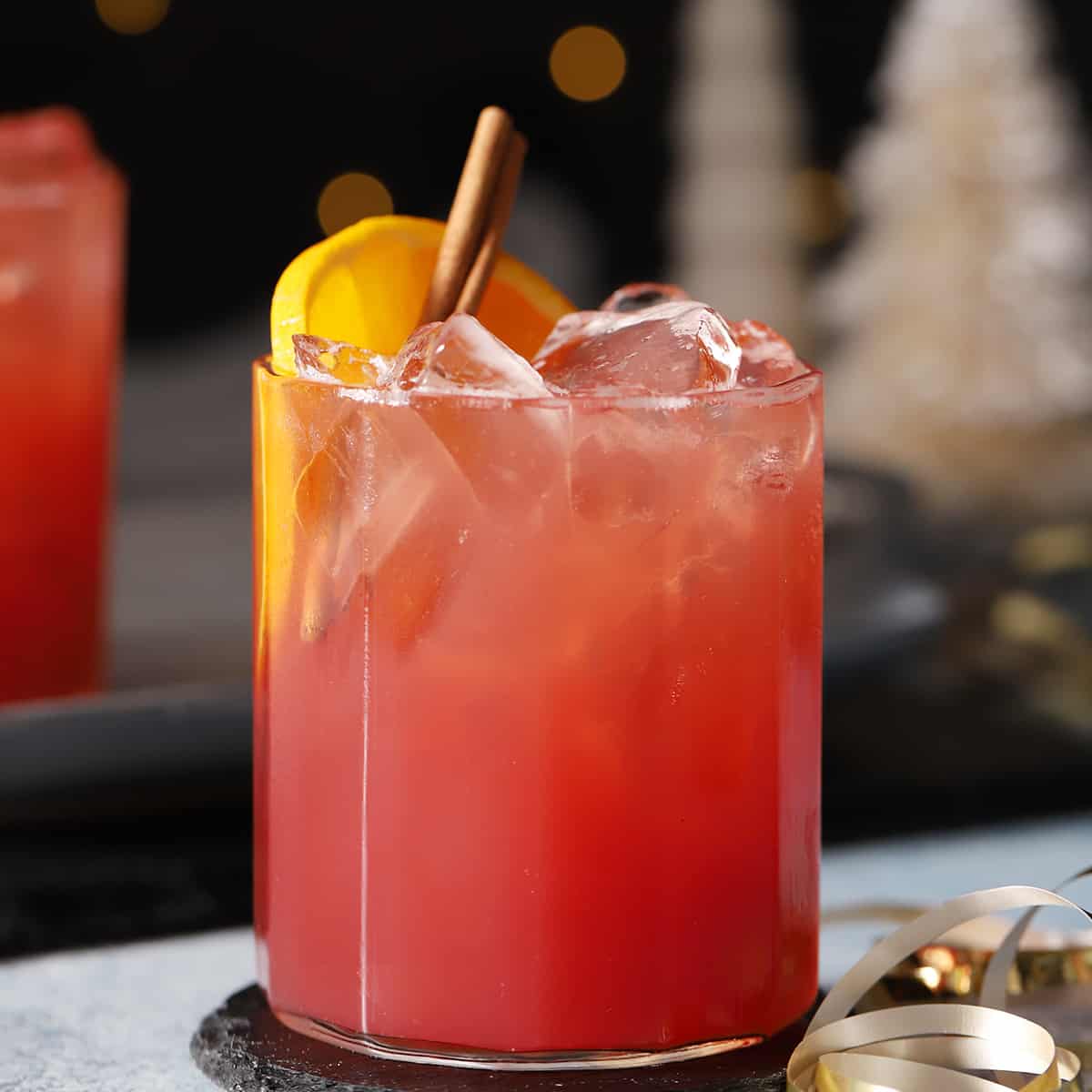 A glass of red-orange cocktail with ice, garnished with an orange slice and a cinnamon stick, sits on a dark coaster with blurred festive decorations in the background.