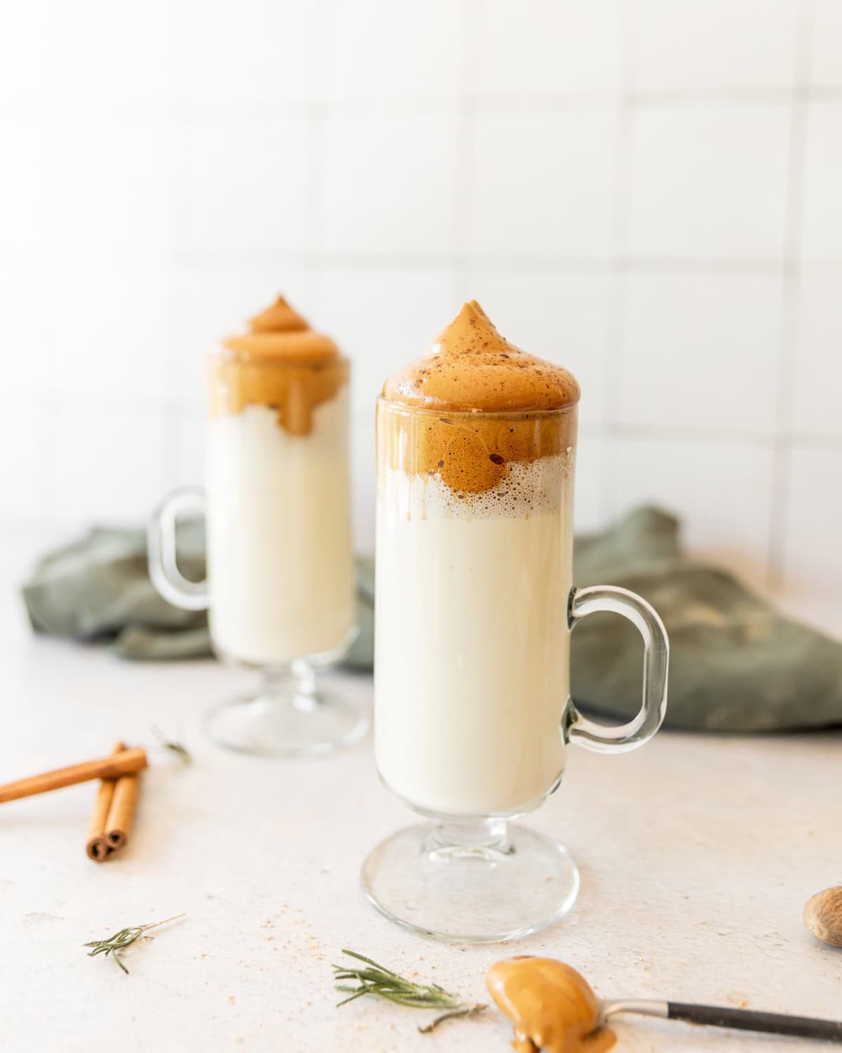 Two glass mugs filled with whipped coffee foam on top of milk, placed on a light surface with cinnamon sticks and herbs nearby.