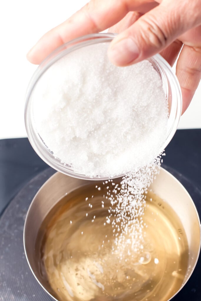 A hand pours granulated sugar from a clear bowl into a pot filled with liquid.