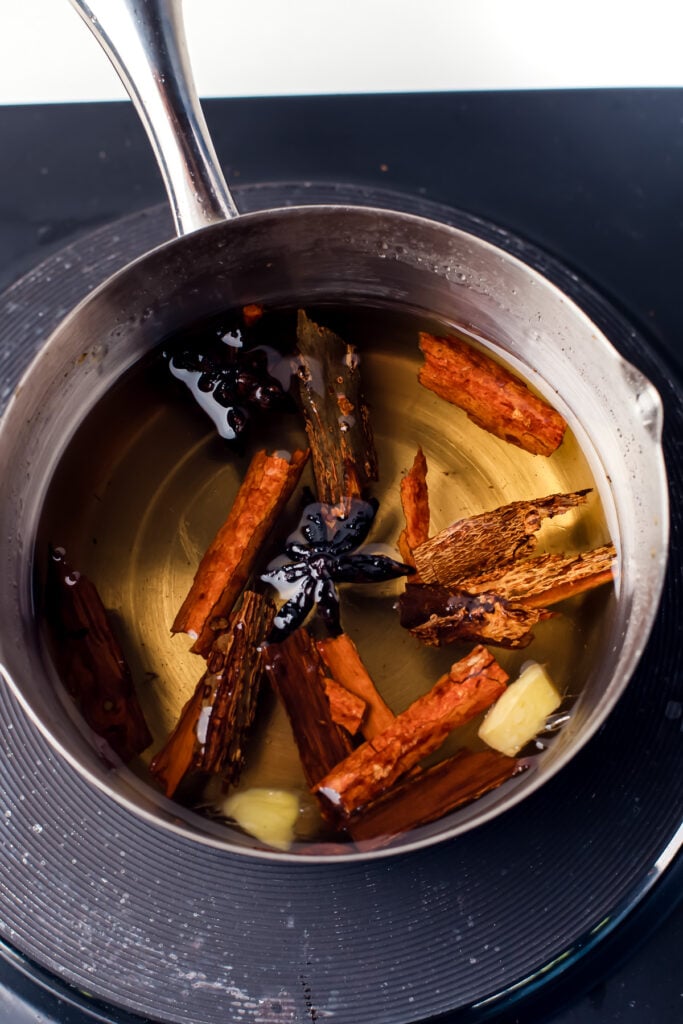 A metal saucepan containing cinnamon sticks, star anise, and ginger simmering in liquid on a stovetop.