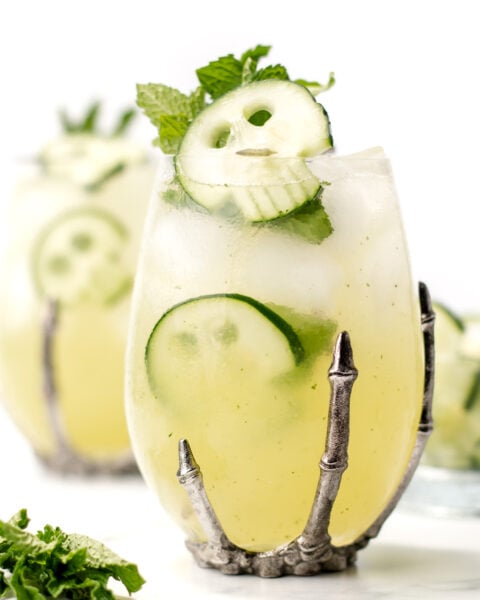 A cocktail garnished with cucumber slices and mint is served in a clear glass with a metal claw-shaped holder, with another similar drink in the background.