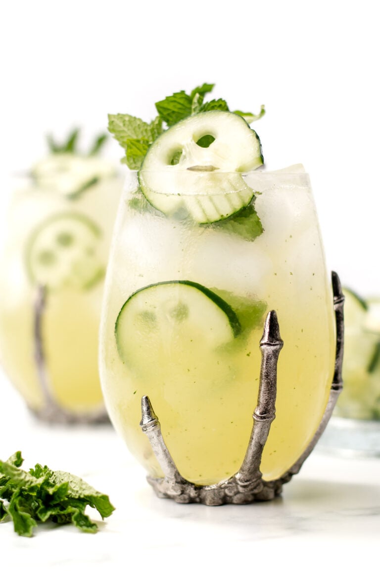 A cocktail garnished with cucumber slices and mint is served in a clear glass with a metal claw-shaped holder, with another similar drink in the background.