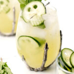 A glass with a metal skeleton hand holder contains an iced cucumber cocktail garnished with mint and cucumber slices. Another drink and mint leaves are in the background.