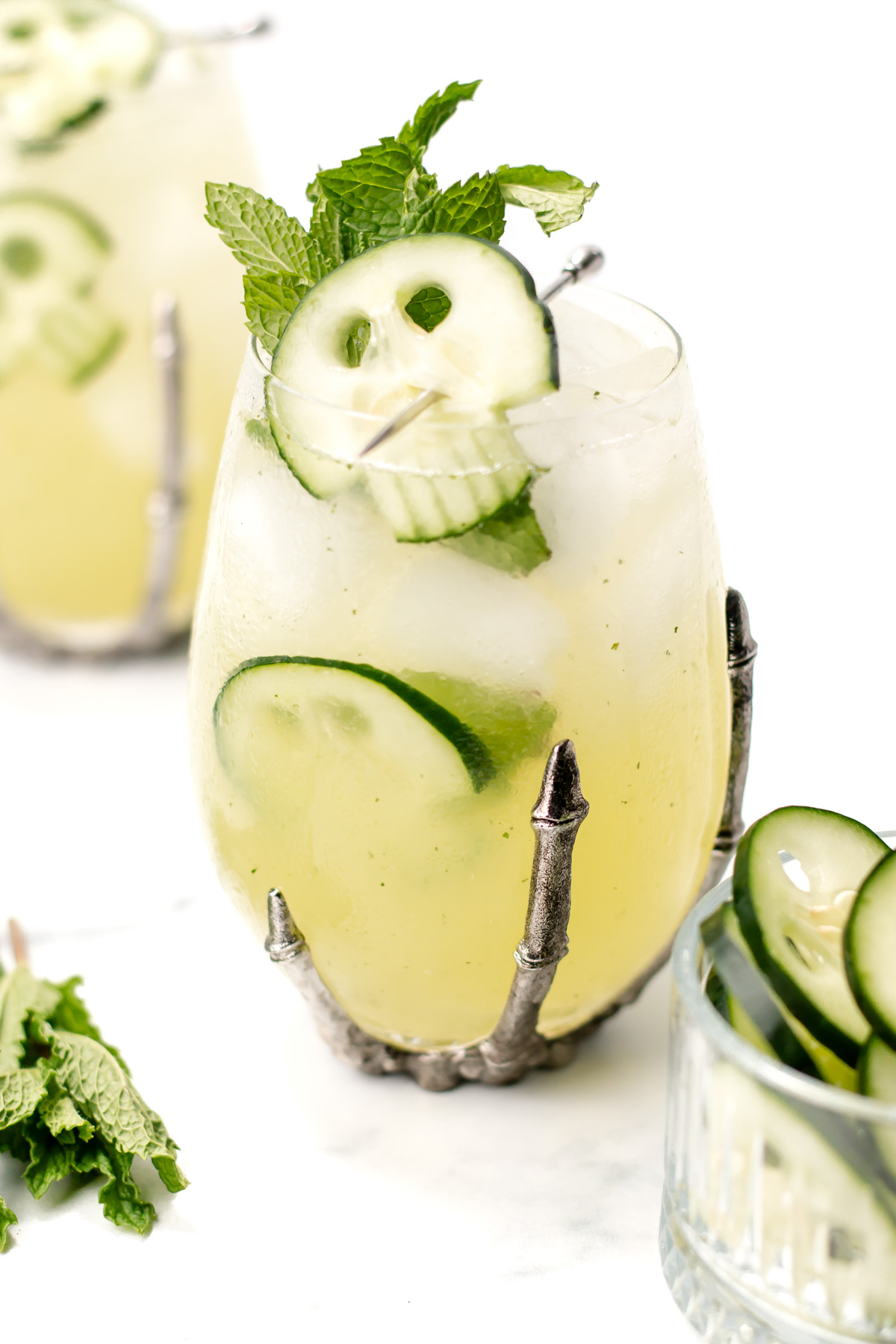 A glass with a metal skeleton hand holder contains an iced cucumber cocktail garnished with mint and cucumber slices. Another drink and mint leaves are in the background.