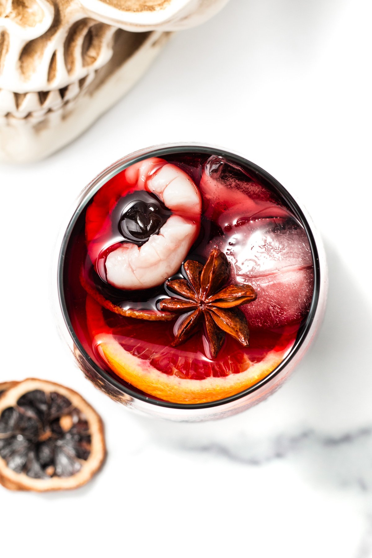 A glass of red punch with ice, a blood orange slice, star anise, and a peeled lychee with a cherry inside, next to a dried citrus slice and part of a skull decoration.