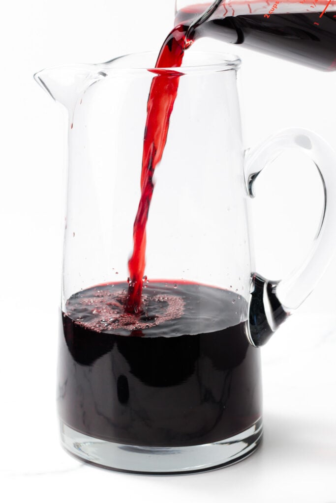 Red liquid being poured from a measuring cup into a clear glass pitcher, partially filled, on a white background.