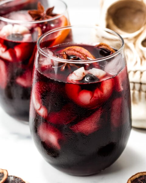 A glass of dark red beverage with ice cubes, garnished with dried fruit and star anise, placed beside a skull decoration.