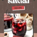 A glass of dark red Halloween sangria with ice and fruit, set in front of a skull decoration and a pitcher of more sangria.