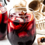 A glass of dark red cocktail with ice, lychee, orange slices, and a star anise garnish, set in front of a realistic skull decoration.