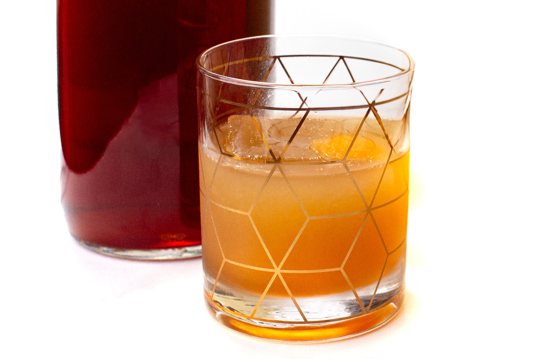 A glass with geometric patterns holds an orange-colored drink with ice, positioned in front of a dark amber liquid in a bottle.