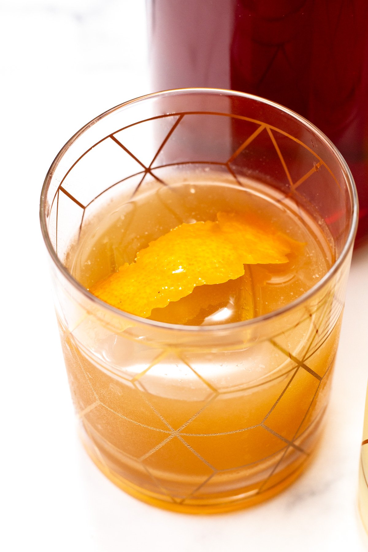 A glass of amber-colored cocktail made with zero-proof whiskey and garnished with an orange peel, served in a patterned tumbler on a white surface.