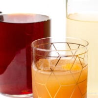 A glass with an amber cocktail and ice sits in front of two glass bottles containing dark and pale liquids.