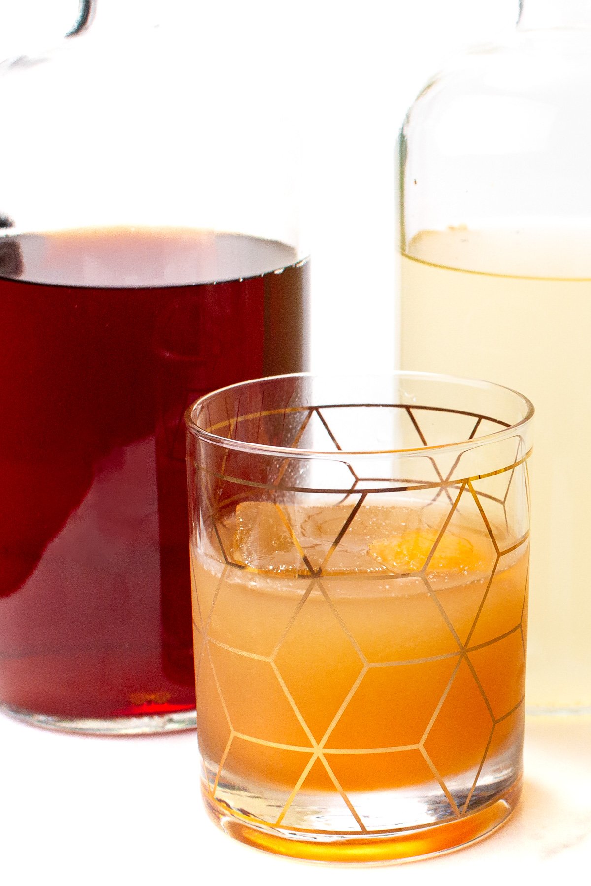 A glass with an amber cocktail and ice sits in front of two glass bottles containing dark and pale liquids.