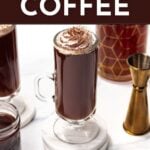 A glass mug of Irish coffee topped with whipped cream and chocolate shavings, surrounded by coffee-related items and bar tools. Text reads "Irish Coffee - Get the Recipe - feastandwest.com".