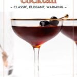 A Manhattan cocktail in a coupe glass, garnished with two dark cherries on a cocktail pick; another glass is partially visible in the background.