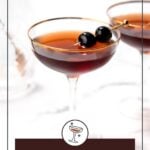 A Manhattan cocktail garnished with two dark cherries on a pick, served in a coupe glass, with another similar glass blurred in the background.