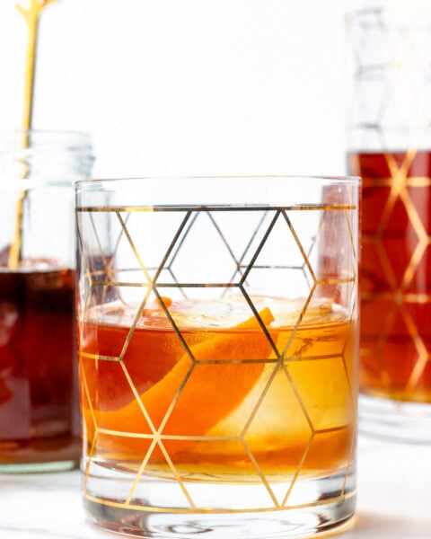 A glass of amber-colored cocktail with ice and an orange peel, featuring a geometric gold pattern, sits on a white surface next to a jar and a decanter.