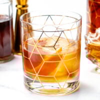 A glass of amber-colored cocktail with large ice cubes, decorated with a geometric gold pattern, sits on a white surface surrounded by bottles of spirits.
