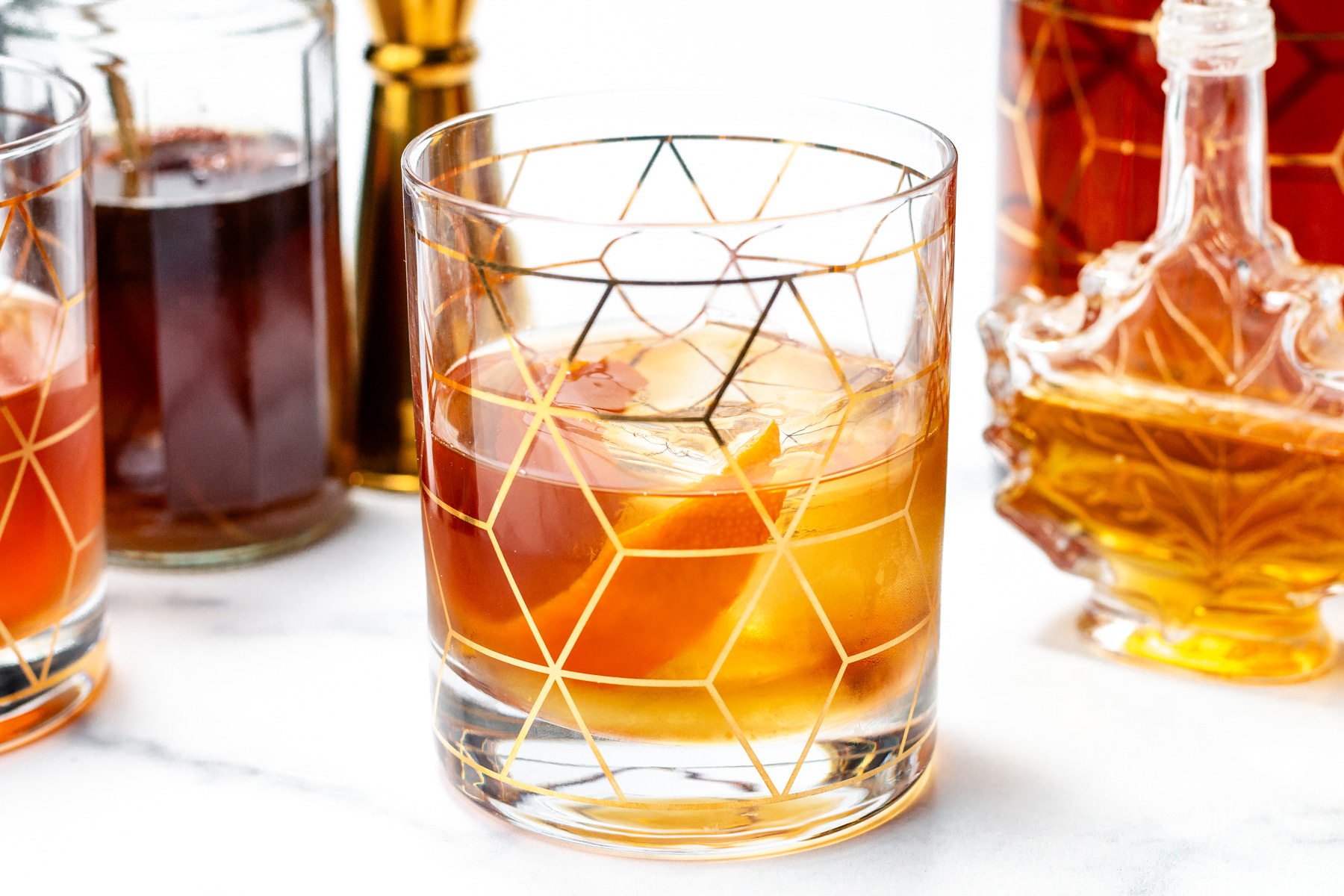 A glass of amber-colored cocktail with large ice cubes, decorated with a geometric gold pattern, sits on a white surface surrounded by bottles of spirits.