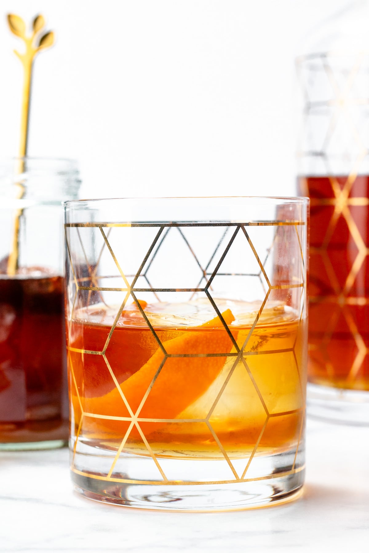 A glass of amber-colored cocktail with ice and an orange peel, featuring a geometric gold pattern, sits on a white surface next to a jar and a decanter.
