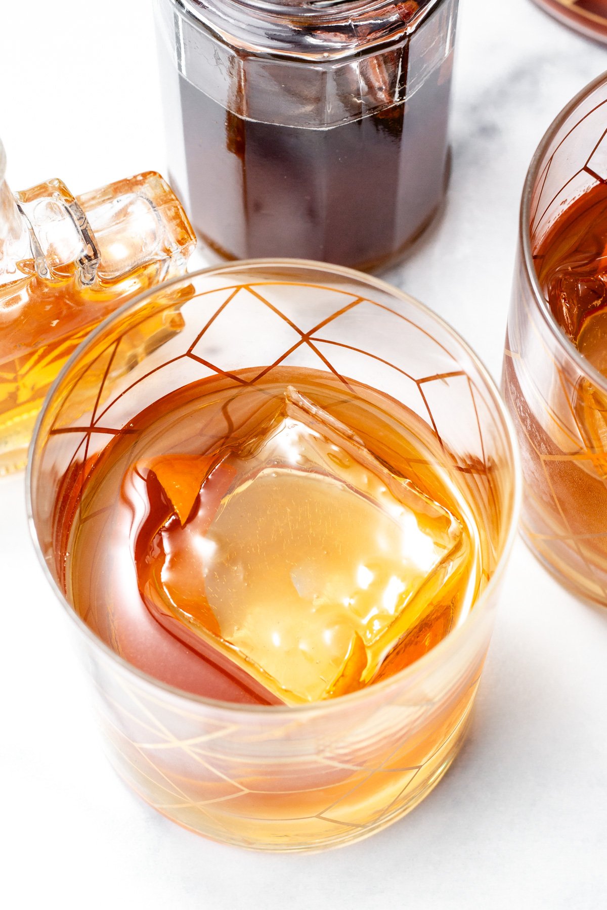 A glass of whiskey with a large square ice cube sits on a white surface, surrounded by bottles containing amber and dark liquids.