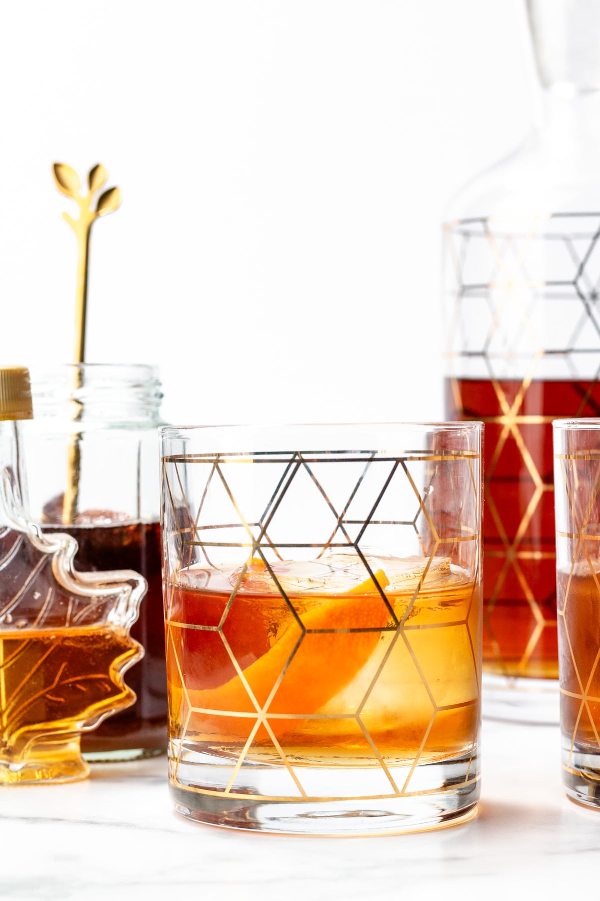A glass of amber cocktail with an orange peel, geometric-patterned glassware, a maple leaf-shaped syrup jar, and a decanter in the background on a white surface.
