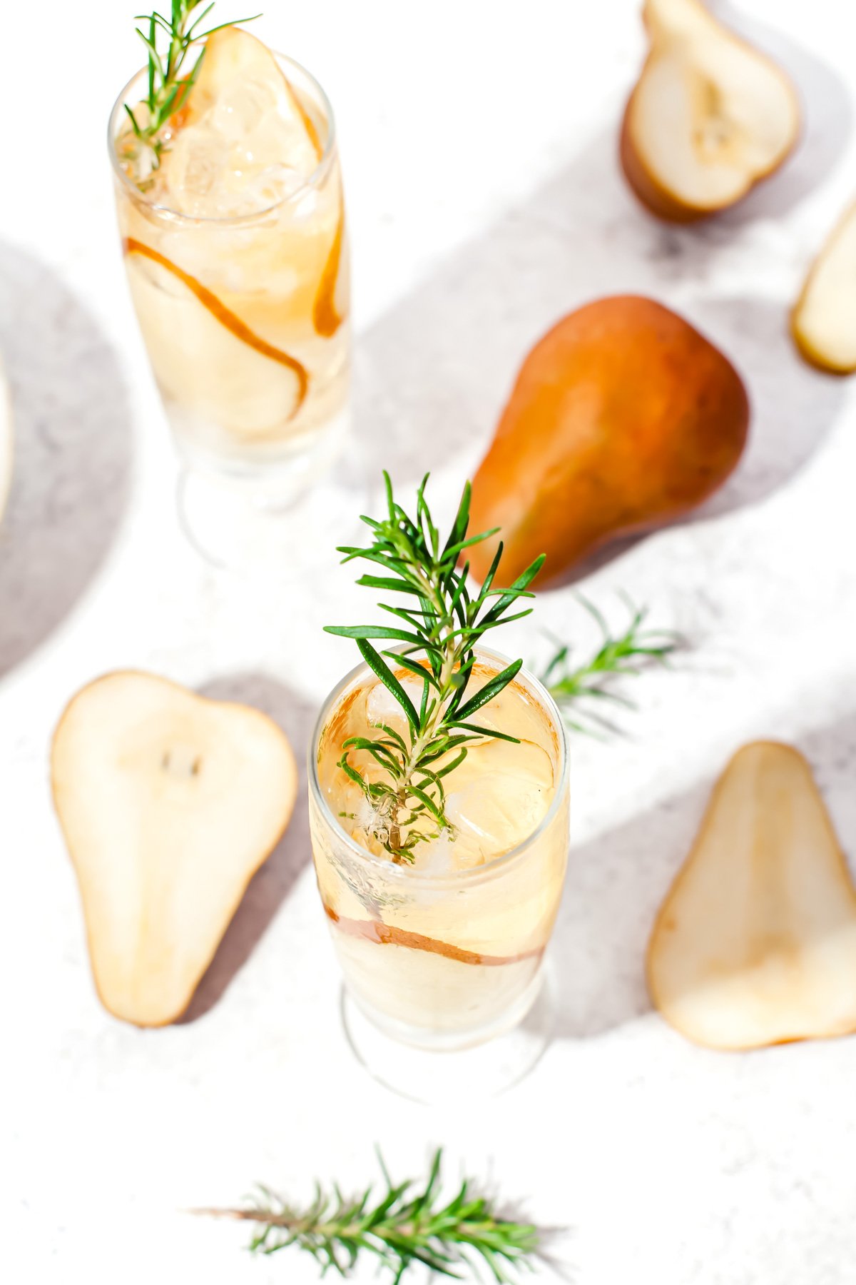 Two glasses of a light-colored drink garnished with rosemary and pear slices, surrounded by pear halves and sprigs of rosemary on a white surface.