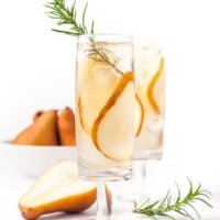 Two glasses of a clear drink garnished with pear slices and rosemary, with a sliced pear and a bowl of whole pears in the background.