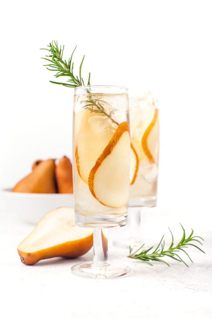 Two glasses of a clear drink garnished with pear slices and rosemary, with a sliced pear and a bowl of whole pears in the background.