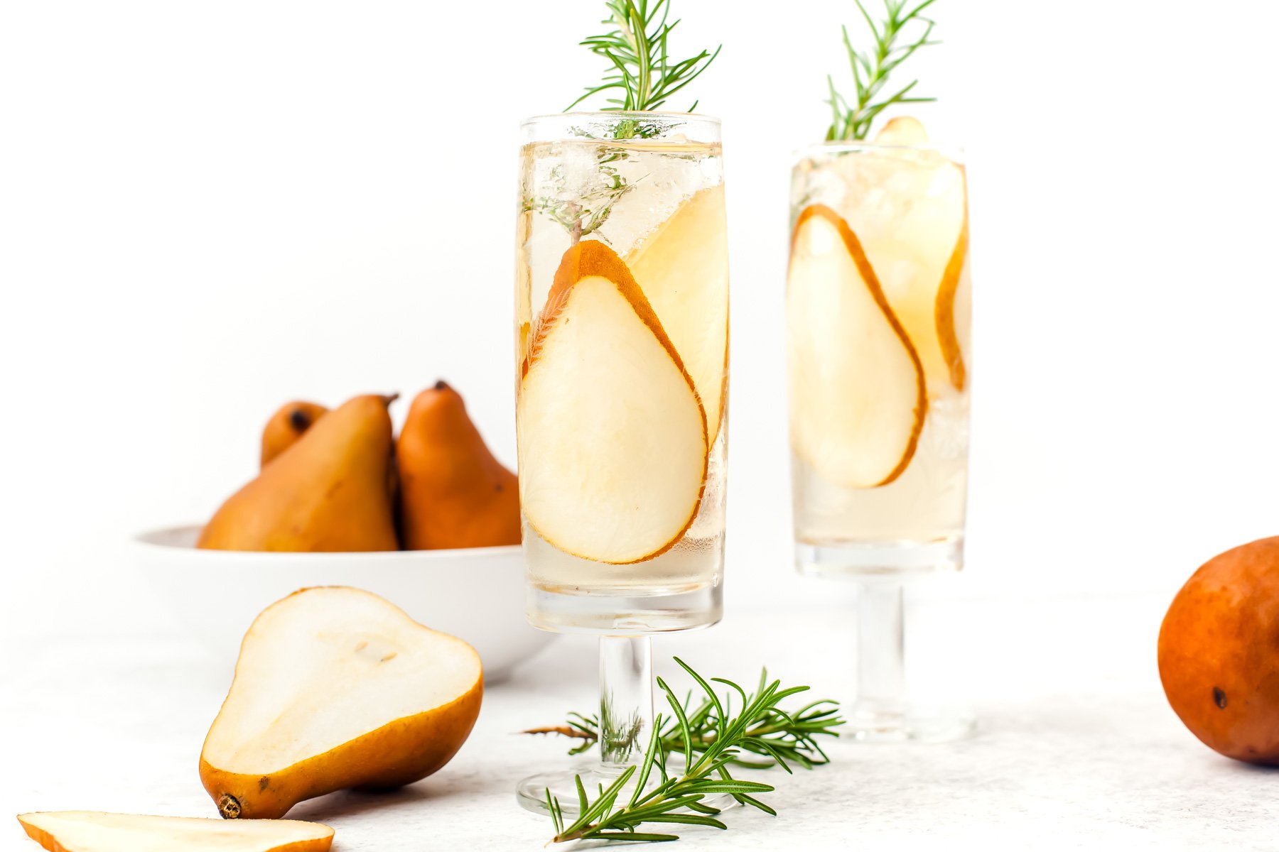 Two glasses of pear cocktails with rosemary and ice, garnished with pear slices, sit on a white surface near a bowl of whole pears and loose pear pieces.