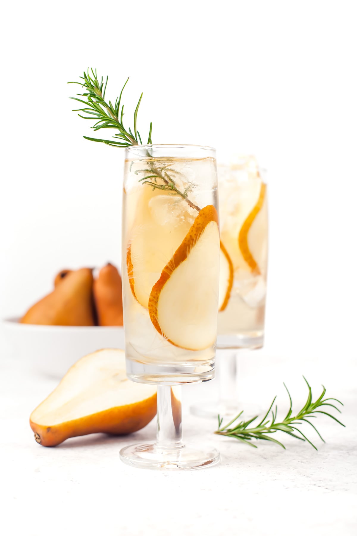 Two glasses of a clear drink garnished with pear slices and rosemary, with a sliced pear and a bowl of whole pears in the background.
