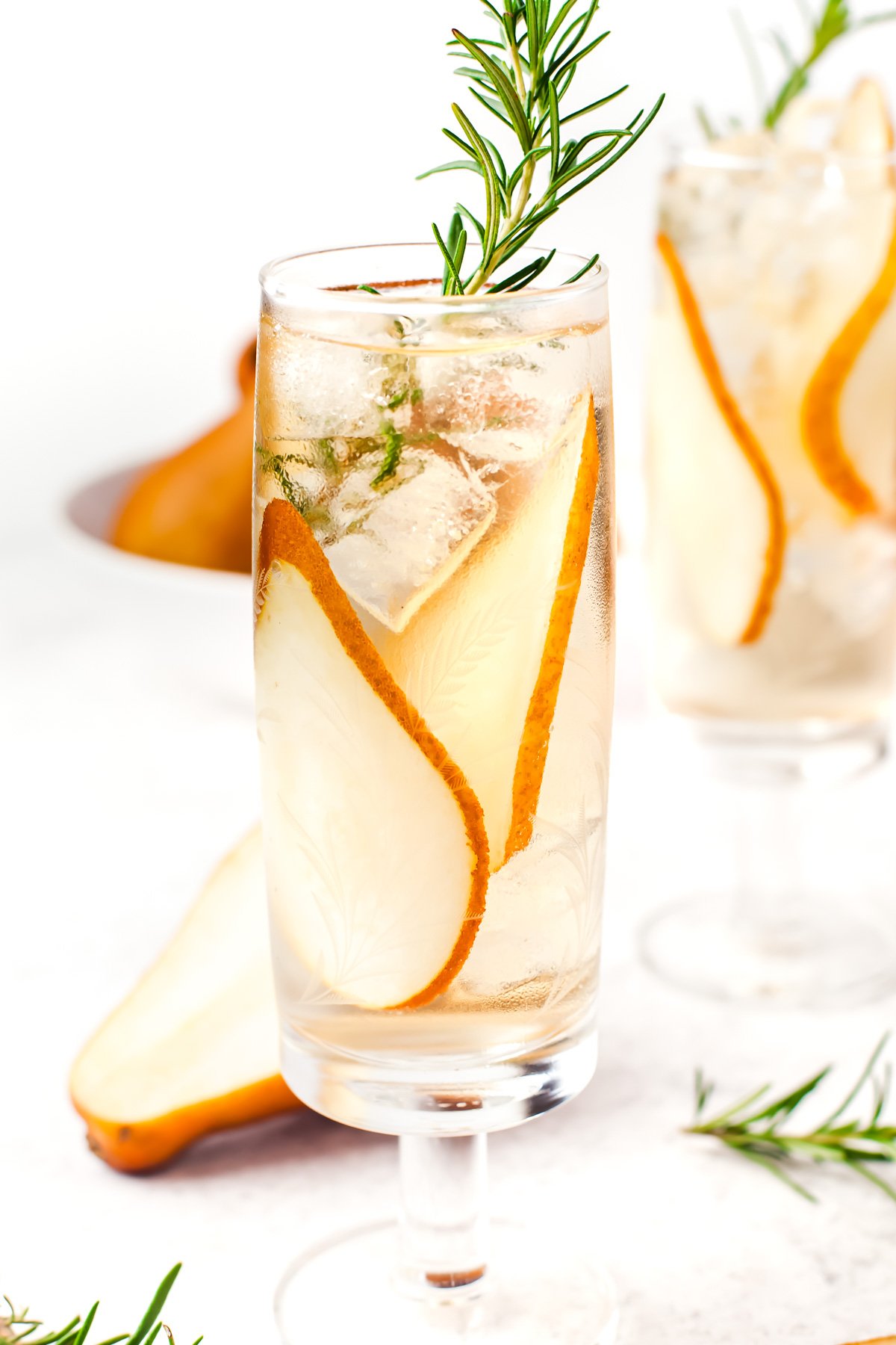 A tall glass filled with ice, clear liquid, pear slices, and garnished with a sprig of rosemary. Another similar glass and a pear are visible in the background.