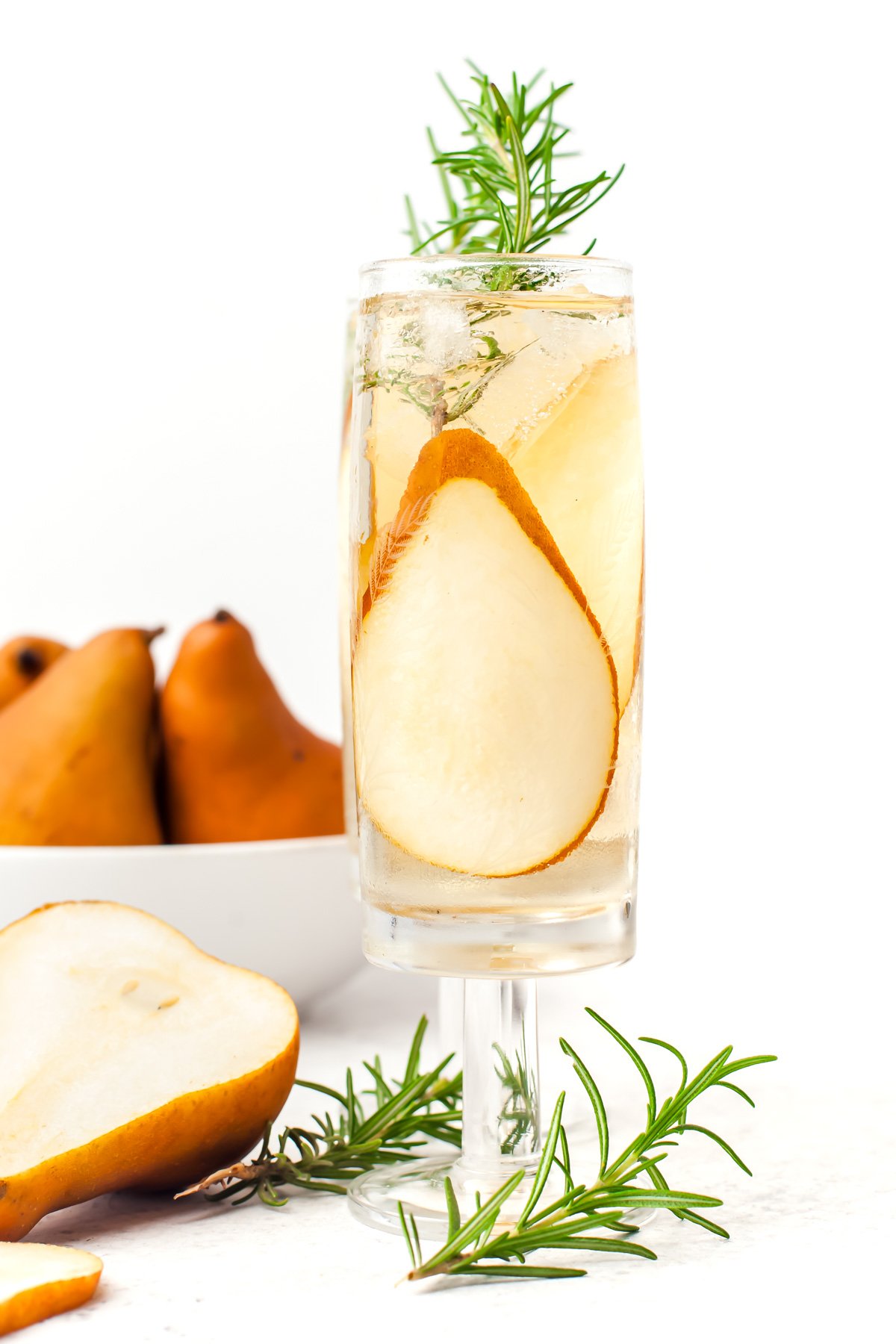 A glass of clear beverage garnished with rosemary and pear slices, with whole pears and more rosemary in the background.