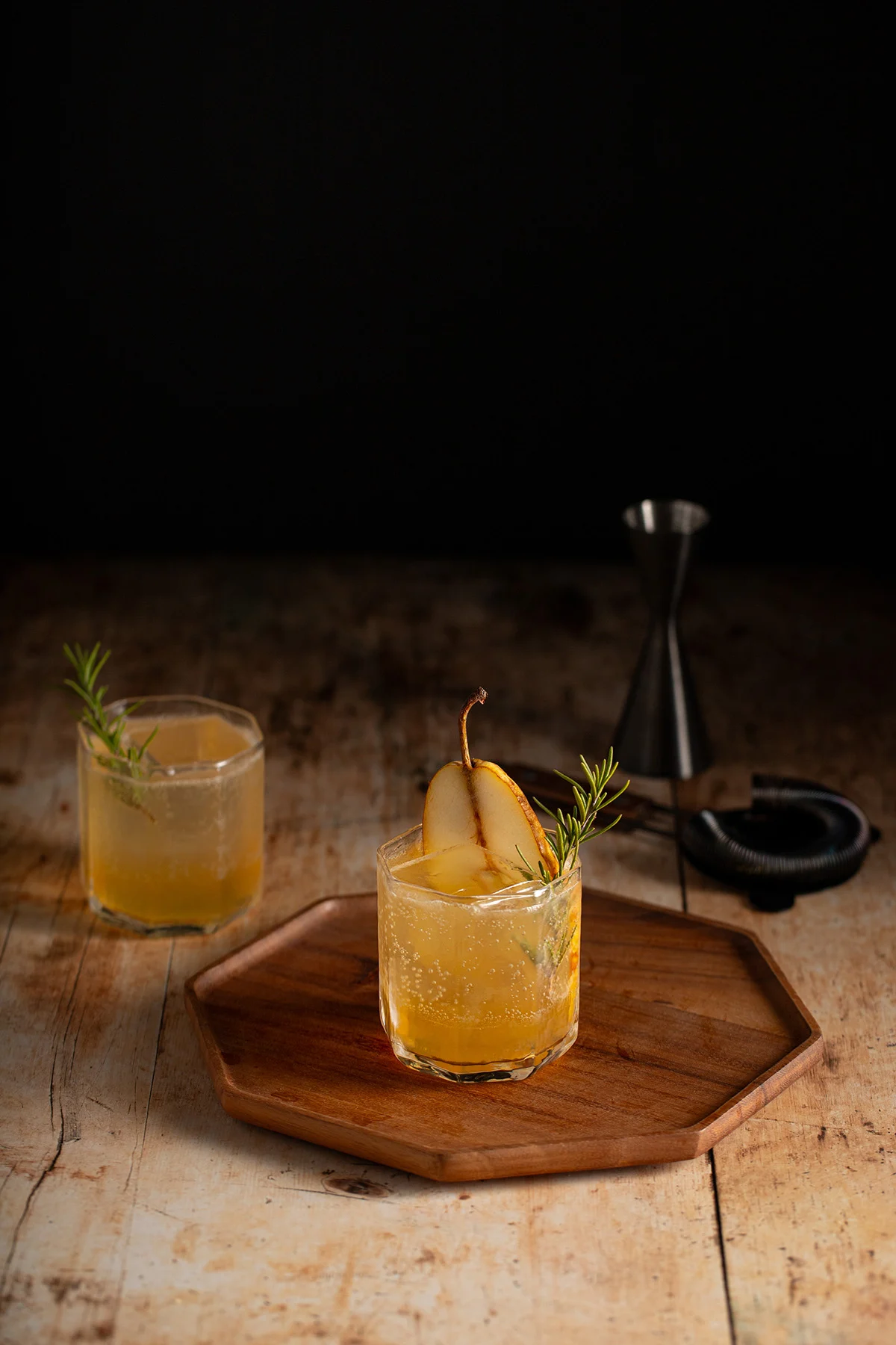 Two Thanksgiving mocktails garnished with pear slices and rosemary sit on a rustic wooden surface—one on a wooden tray—with a cocktail jigger and bar tool in the background.